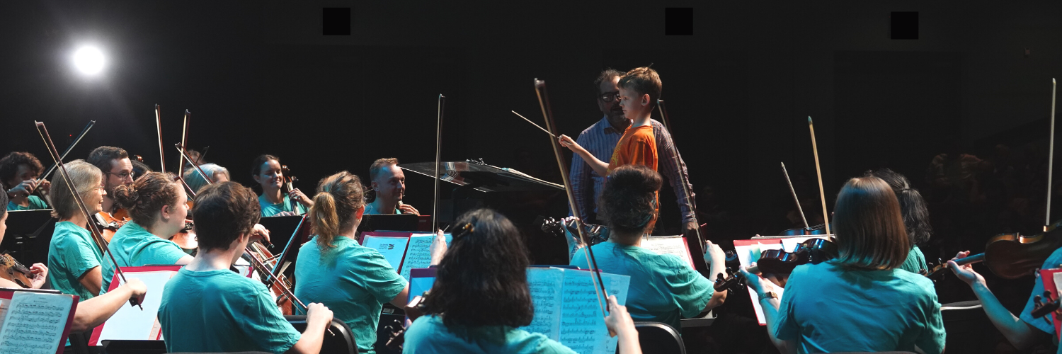 Young boy conducting the Darwin Symphony Orchestra during the orchestra's 2021 performance of Family Proms. Image includes many musicians facing the young boy playing string instruments